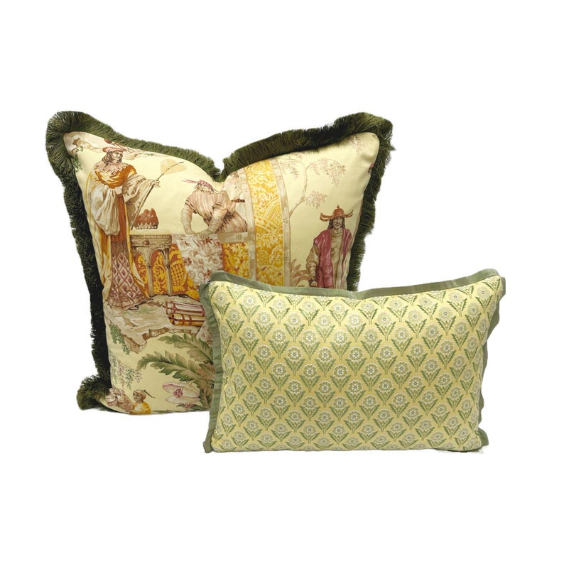 May include: Two decorative throw pillows with a green fringe. The larger pillow has a cream background with a colorful floral pattern. The smaller pillow has a yellow background with a green geometric pattern.