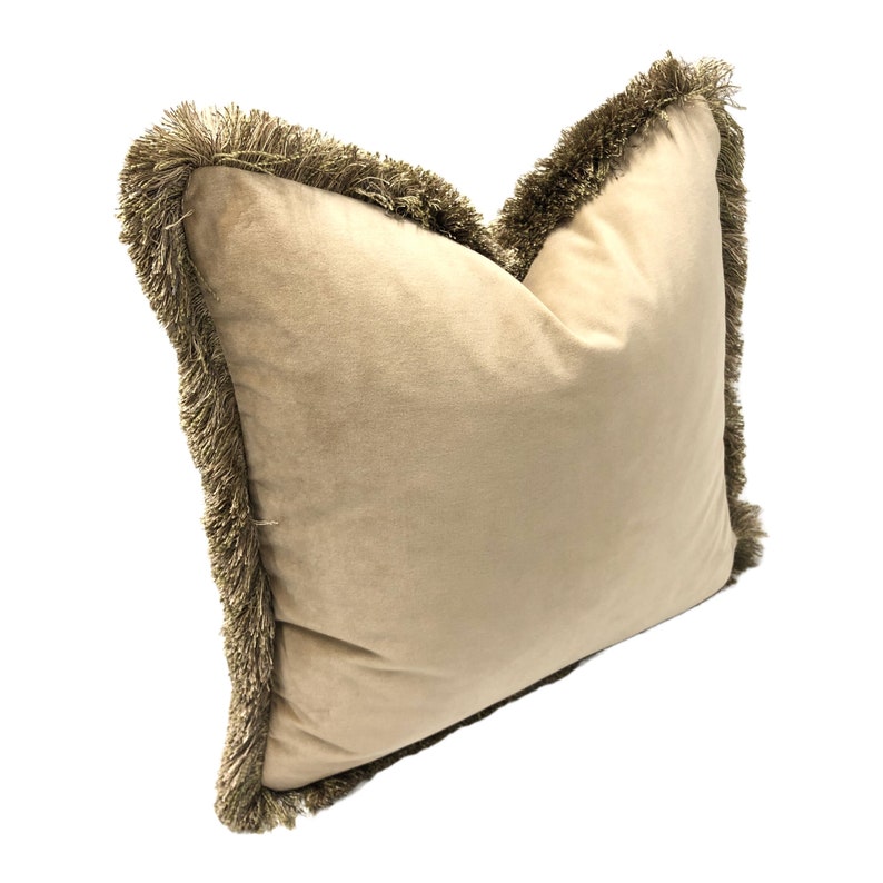 May include: A beige velvet pillow with a brown fringe trim around the edges.