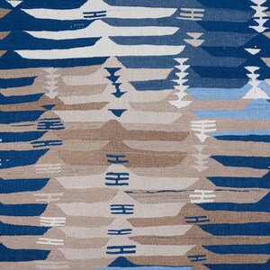 May include: A woven textile with a repeating geometric pattern. The design features horizontal bands of blue, white, and tan, creating a layered effect. The pattern includes stylized mountain-like shapes and linear accents.