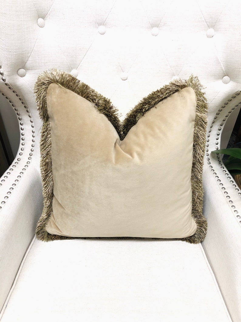 May include: A beige velvet pillow with a brown fringe trim. The pillow is sitting on a white chair.