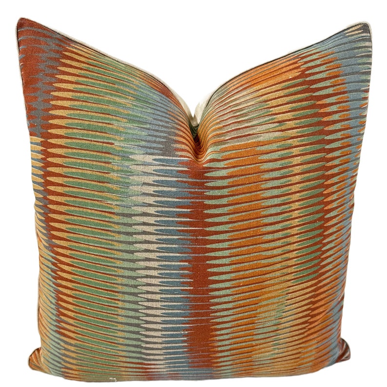 Puede incluir: Una almohada decorativa con un patr&oacute;n geom&eacute;trico en tonos naranja, verde, azul y marr&oacute;n. La almohada tiene un brillo sutil y una tela suave y texturizada.