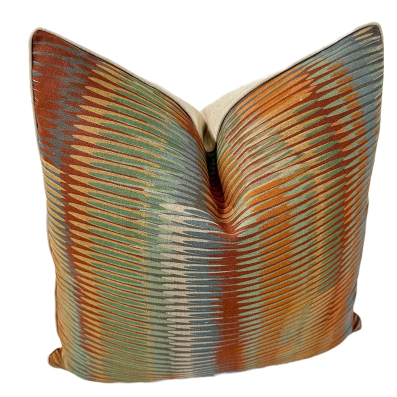 Puede incluir: Una almohada decorativa con un patr&oacute;n geom&eacute;trico en tonos naranja, marr&oacute;n, verde y azul. La almohada tiene un respaldo de lino beige.