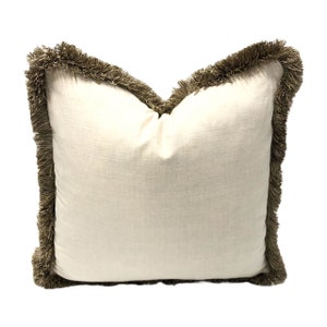 May include: A white pillow with a brown fringe border. The pillow is made of a soft, textured fabric.