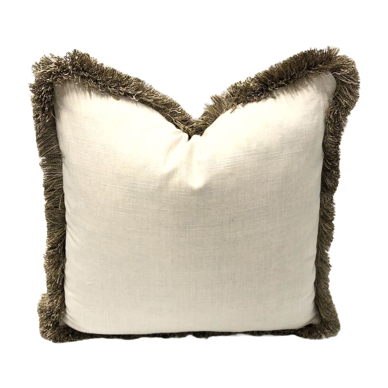 May include: A white pillow with a brown fringe border. The pillow is made of a soft, textured fabric.