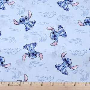 May include: Pale blue fabric with a repeating pattern of the character Stitch from Lilo & Stitch. The design includes Stitch in various poses, along with stylized waves and stars. The fabric is marked with a ruler in inches.