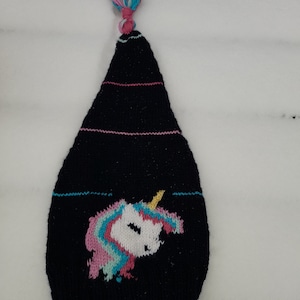 Può includere: Cappello di lana nero con un designo di unicorno e un pon pon di lana rosa, blu e bianco.