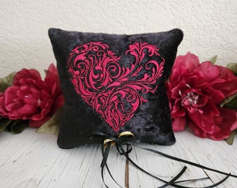 May include: Black velvet ring pillow with a large embroidered red heart design. Two gold wedding rings are attached with a black ribbon. Red flowers are in the background. A decorative item for a wedding.