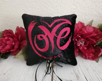 May include: A black ring pillow with the word "Love" embroidered in pink. The pillow is square-shaped and has a textured surface. Black ribbons and a gold bell are attached to the bottom. Red flowers are in the background.