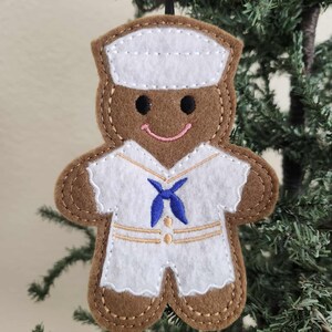 May include: A felt gingerbread man Christmas ornament. The ornament is brown with a white sailor uniform, a blue necktie, and a white hat. The gingerbread man has a pink smile and black eyes. The ornament is hanging on a black string.