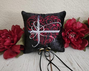 May include: Black velvet ring pillow with a red embroidered heart design and white accents. The heart features a cross and swirling patterns. Two gold rings are attached with black ribbons. Red flowers are on either side.