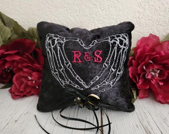 May include: Black ring pillow with a heart-shaped design made of skeleton hands embroidered in white. The initials "R&S" are embroidered in pink. Two gold rings are attached with a black ribbon. The pillow is surrounded by red flowers.