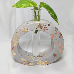 Puede incluir: Un jarrón de hormigón gris con detalles de pan de oro y un tubo de ensayo de vidrio transparente en el centro. Una pequeña planta verde está en el tubo de ensayo.