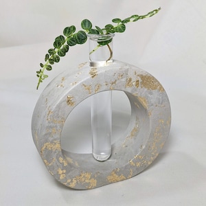 Puede incluir: Un jarrón de hormigón gris con detalles dorados, con forma de anillo, con un tubo de ensayo de vidrio en el centro. Una pequeña planta verde está en el tubo de ensayo.