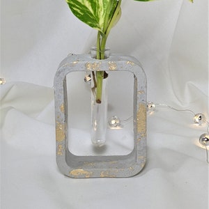 Puede incluir: Un jarrón de hormigón gris con detalles dorados contiene un solo tallo de una planta variegada con hojas verdes y amarillas. El jarrón tiene un tubo de ensayo de vidrio transparente en el centro.