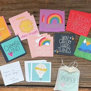 May include: A collection of colorful square cards with inspirational quotes and illustrations, including rainbows, suns, and mountains. The cards are arranged on a wooden surface, with a small drawstring bag in the corner. Text on the cards includes "Dream Big Dreams".