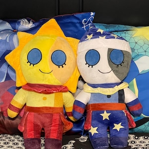 Puede incluir: Dos peluches, uno con forma de sol con una cara amarilla y naranja y ropa roja y naranja, el otro con forma de luna con una cara blanca y gris y ropa azul y blanca.