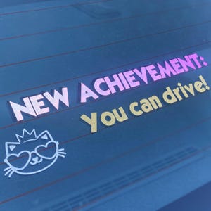 May include: A vinyl decal applied to a car window featuring the text NEW ACHIEVEMENT: in a pink gradient font and You can drive! in a yellow font. Below the text is a white line art illustration of a cat wearing a crown and heart-shaped glasses.