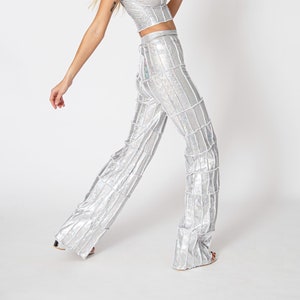 Op de afbeelding: Een zilveren twee-delige outfit met een getailleerd topje en een wijde broek. De stof heeft een glinsterende, iriserende afwerking en is in verticale panelen gesneden.