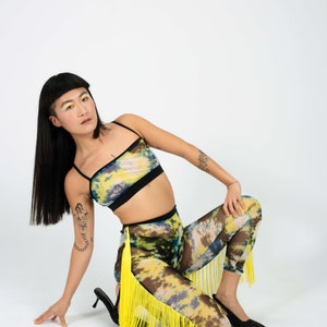 Op de afbeelding: Een vrouw draagt een geel en blauw tie-dye crop top en bijpassende leggings met gele franjes. Ze poseert op een witte achtergrond.