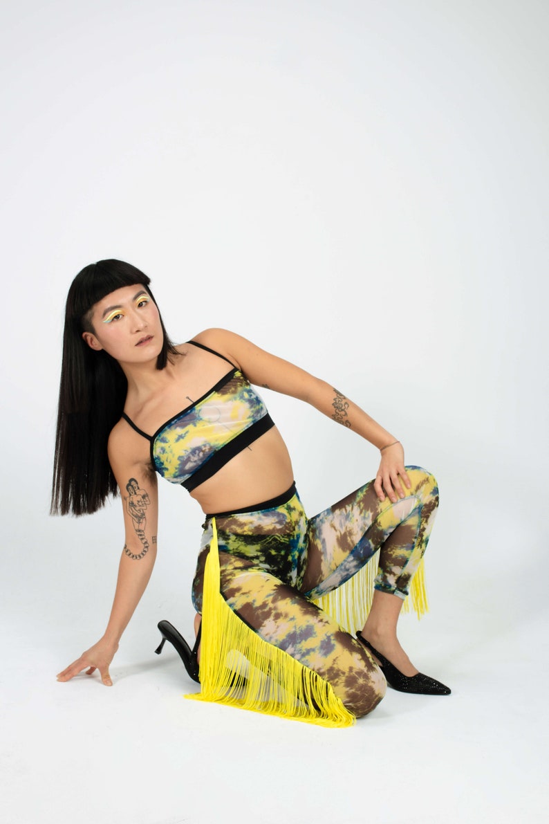 Op de afbeelding: Een vrouw draagt een geel en blauw tie-dye crop top en bijpassende leggings met gele franjes. Ze poseert op een witte achtergrond.