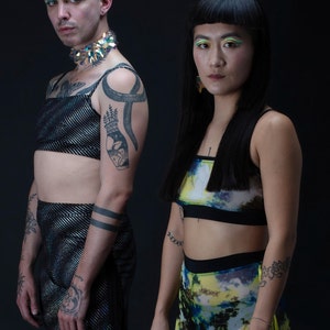 Op de afbeelding: Twee mensen dragen kleurrijke kleding en accessoires. De persoon links draagt een zwart en zilveren paillettentop, een zwarte broek en een zilveren en turquoise choker. De persoon rechts draagt een zwart en blauw crop top, een gele en blauwe geprinte broek en een paar oorbellen.