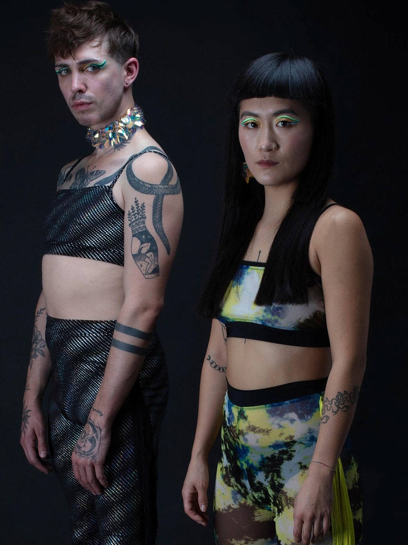 Op de afbeelding: Twee mensen dragen kleurrijke kleding en accessoires. De persoon links draagt een zwart en zilveren paillettentop, een zwarte broek en een zilveren en turquoise choker. De persoon rechts draagt een zwart en blauw crop top, een gele en blauwe geprinte broek en een paar oorbellen.