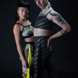 Op de afbeelding: Twee mensen dragen kleurrijke kleding en accessoires. De persoon links draagt een top met een groen, geel en blauw patroon met een gele franje. De persoon rechts draagt een zilveren en zwarte top met een zwarte broek. Beide mensen hebben kleurrijke make-up.