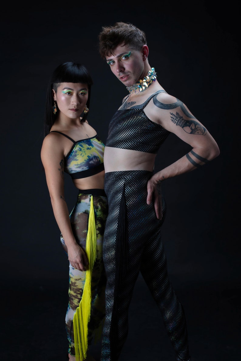 Op de afbeelding: Twee mensen dragen kleurrijke kleding en accessoires. De persoon links draagt een top met een groen, geel en blauw patroon met een gele franje. De persoon rechts draagt een zilveren en zwarte top met een zwarte broek. Beide mensen hebben kleurrijke make-up.