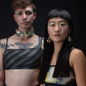 Op de afbeelding: Twee mensen dragen zwarte tops met kleurrijke accenten. De persoon links draagt een zwarte top met zilveren glitter en een glinsterende groen-blauwe choker. De persoon rechts draagt een zwarte top met een kleurrijke abstracte print. Beide mensen hebben kleurrijk make-up rond hun ogen.