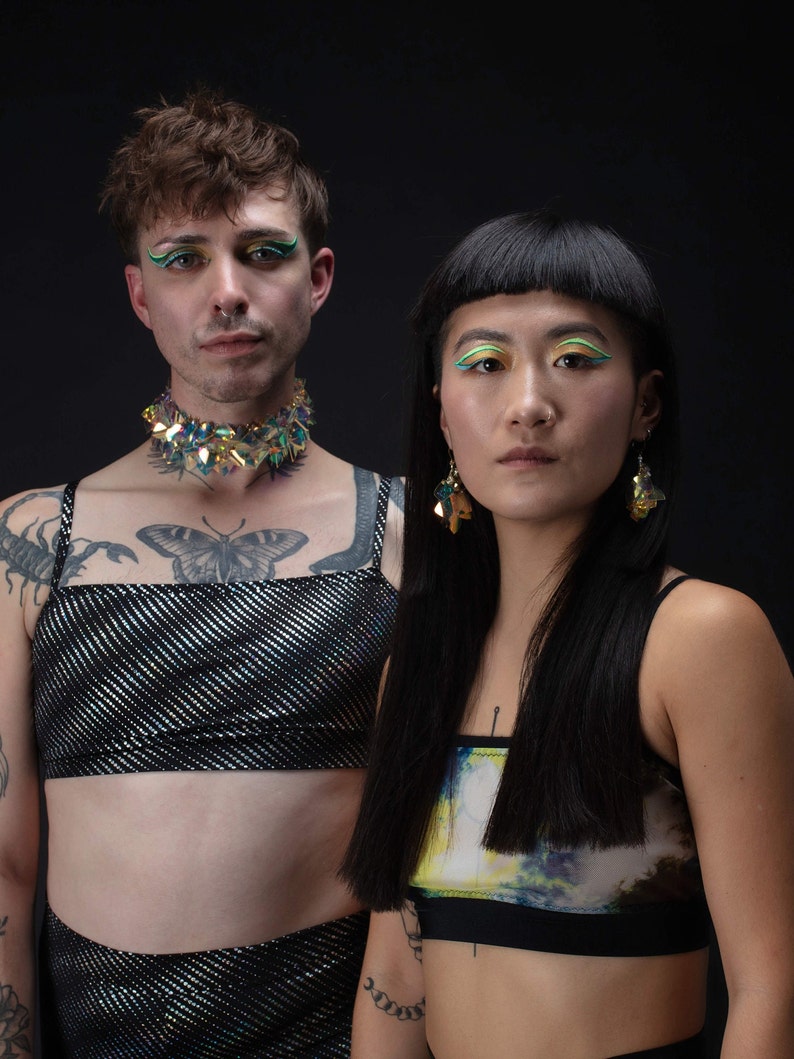 Op de afbeelding: Twee mensen dragen zwarte tops met kleurrijke accenten. De persoon links draagt een zwarte top met zilveren glitter en een glinsterende groen-blauwe choker. De persoon rechts draagt een zwarte top met een kleurrijke abstracte print. Beide mensen hebben kleurrijk make-up rond hun ogen.