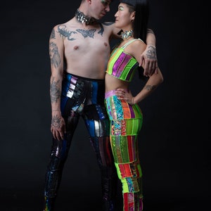 May include: Two people wearing colorful patchwork pants and tops. The person on the left is wearing a silver necklace and has tattoos on their arms. The person on the right is wearing a green and pink top and a silver necklace. They are looking at each other.