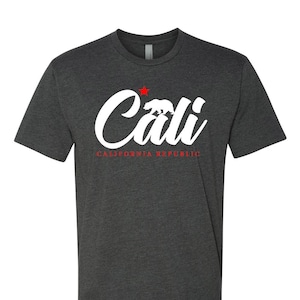 May include: A dark heather grey t-shirt with a white graphic of the word "Cali" in a stylized font. A red star and a white silhouette of a bear are above the word. The text "CALIFORNIA REPUBLIC" is written in red below the graphic.