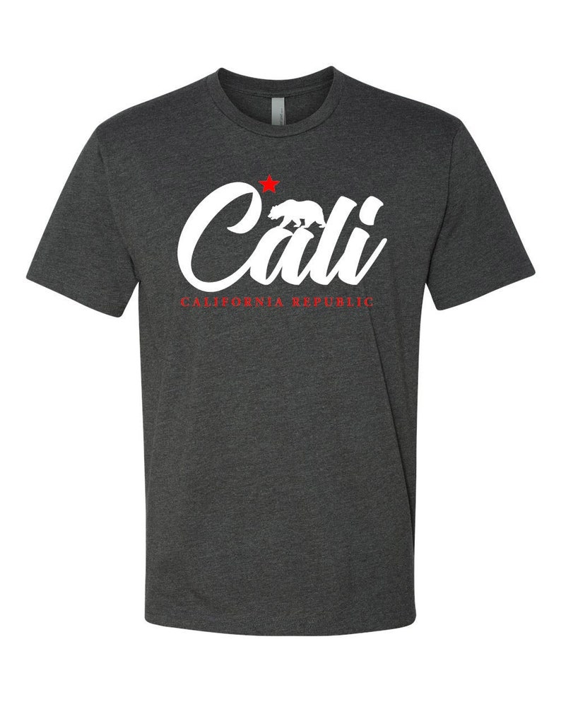 May include: A dark heather grey t-shirt with a white graphic of the word "Cali" in a stylized font. A red star and a white silhouette of a bear are above the word. The text "CALIFORNIA REPUBLIC" is written in red below the graphic.
