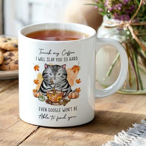 White ceramic coffee mug with a humorous cat design. The mug features a grumpy grey tabby cat with a coffee cup, surrounded by autumn leaves and pumpkins. Text on the mug reads "Touch my Coffee I WILL SLAP YOU SO HARD, EVEN GOOGLE WON'T BE Able to find you."