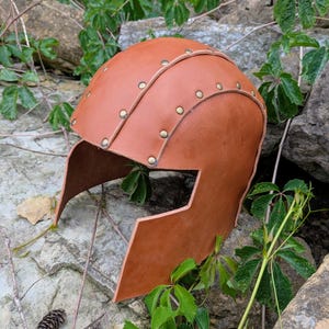 May include: A tan leather helmet featuring a rounded crown and a face-covering design. The helmet is constructed from multiple overlapping leather panels secured together with small, evenly spaced metallic rivets along the seams.
