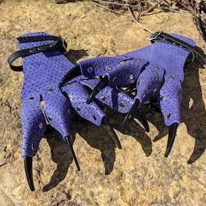 Könnte beinhalten: Ein Paar lila Krallengloves mit schwarzen Krallen und Handgelenksriemen. Die Handschuhe haben eine schuppenartige Textur und sind mit silbernen Nieten besetzt. Die Krallen sind lang und gebogen.