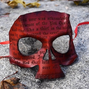 May include: A red skull-shaped mask featuring black printed text from The Masque of the Red Death. The mask has two large eye openings, a triangular nose cutout, and thin red ribbons attached to the sides for securing the item to the face.