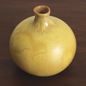May include: A spherical, yellow ceramic vase with a narrow neck. The vase has a smooth, shiny finish and a slightly textured surface. The opening is a darker, natural wood colour. The vase is on a dark brown wooden surface.
