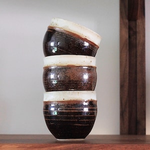 May include: Three brown and white ceramic mugs stacked on top of each other on a wooden shelf.