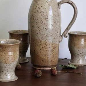 May include: A set of four speckled brown and white ceramic cups and a matching jug. The jug has a wide mouth and a handle. The cups are all the same size and have a rounded shape.