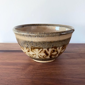 May include: A handcrafted ceramic bowl with a rustic appearance. The bowl has a light brown exterior with darker brown floral patterns around the base. The interior is a darker shade of brown. The bowl is on a wooden surface.