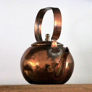 May include: A copper kettle with a rounded body and a large, arched handle. The spout is curved, and a small knob sits atop the lid. The kettle has a polished, reflective surface, showing signs of age and use. It is a vintage kitchenware item.