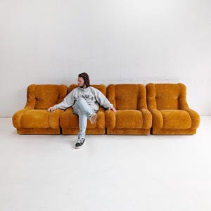 May include: A four-piece, mustard-colored sofa with a rounded design. The sofa has a plush, textured appearance. The image shows a person sitting on the sofa. The sofa is against a white wall.