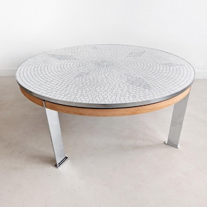 Op de afbeelding: Een ronde salontafel met een mozaïektegelblad en een houten rand. De tafel heeft een diameter van ongeveer 100 cm en staat op vier zilveren metalen poten.