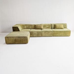 Może przedstawiać: Modułowa oliwkowozielona sofa narożna z pasującym pufem. Sofa ma niski profil, kwadratowy kształt i zawiera kilka kwadratowych poduszek do rzucania. Tkanina ma teksturowany wygląd, a elementy są ustawione na białej ścianie.
