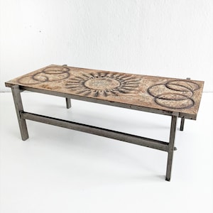 Op de afbeelding: Een rechthoekige salontafel met een metalen frame en een getextureerd blad. Het tafelblad heeft een zonmotief en twee sets in elkaar grijpende cirkels. De tafel wordt ondersteund door vier metalen poten.