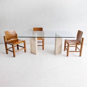 Op de afbeelding: Een moderne eettafel met een helder glazen blad en een unieke stenen voet. Twee houten stoelen met leren zittingen en een houten stoel zijn rond de tafel geplaatst. De tafel is ongeveer 183 cm lang.