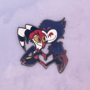 May include: A red and black cartoon character with horns and a bow tie is hugging a blue cartoon character with a long snout. The characters are smiling and have their eyes closed. The pin is made of metal and has a shiny finish.