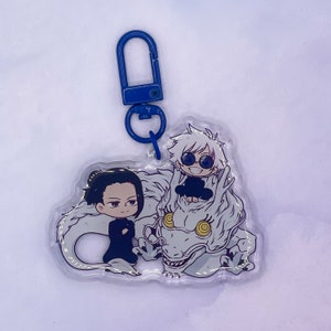 May include: A white dragon keychain with two anime characters. The dragon is white with yellow eyes and a black mouth. One character is a white-haired boy with black sunglasses and a black shirt. The other character is a black-haired girl with a black shirt and black hair.