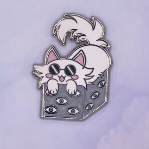 May include: A white cat wearing sunglasses sits inside a gray box with multiple eyes printed on the sides. The cat has pink ears and a white fluffy tail.